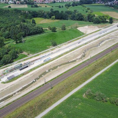 Ein Bach führt neben Bahngleisen und einer Baustelle entlang.