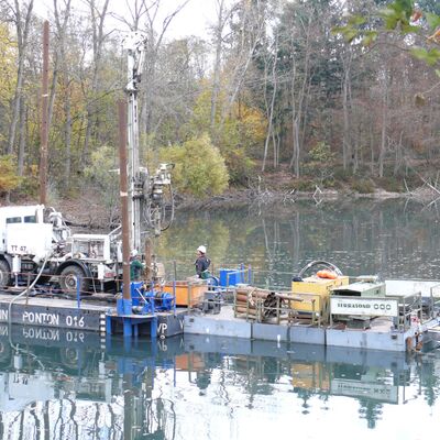 Ein Bohrgerät steht auf einer Plattform mitten auf dem See und wird von zwei Arbeiter:innen gesteuert.