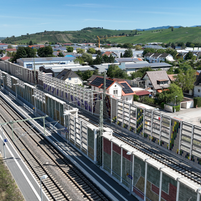 Mehrgleisige Bahnstrecke mit Schallschutzwänden neben einer Straße, im Hintergrund Wohnhäuser, Industriegebäude und hügelige Landschaft.