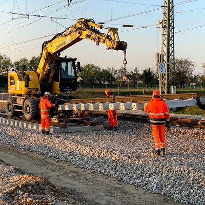Bauarbeiter stehn um eine Baustellenmaschine. Die Maschine hebt ein Gleisteil hoch.