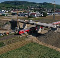 Ein roter Zug fährt unter einer Straßenüberführung durch.
