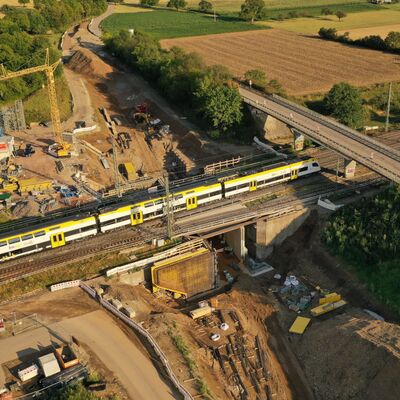 Ein gelber Zug fährt an einer Baustelle unter einen Bahnübergang durch.