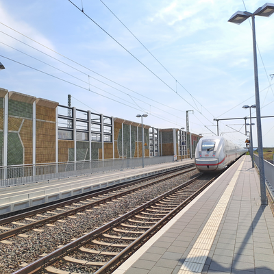 Ein Bahnhof mit einem einfahrenden Zug und einer farbig und regional gestalteten Schallschutzwand im Hintergrund.