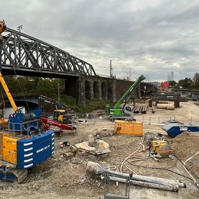 Eine Baumaschine steht auf einer Baustelle. Im Hintergrund ist eine Brücke zu erkennen.