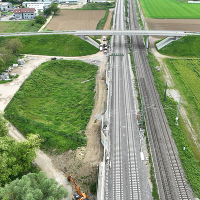 Luftaufnahme einer Straßenüberführung über eine viergleisige Bahnstrecke, umgeben von Feldern, Gebäuden und Baustelleneinrichtungen.