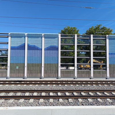 Ein Gleis mit einer Lärmschutzwand im Hintergrund, die mit blauen Bergmotiven gestaltet ist. Sie hat transparente Elemente zum Durchschauen.