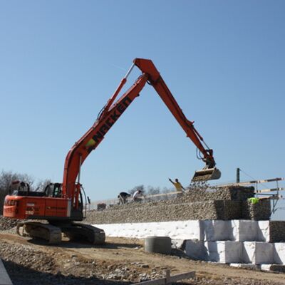 Ein roter Bagger schüttet eine Wand aus Gabionen auf.