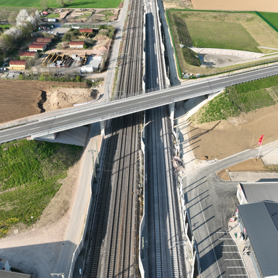 Luftaufnahme einer Straßenüberführung über eine Bahnstrecke, umgeben von Feldern, Gebäuden und Fahrzeugen