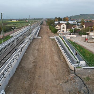 Links die Gleise der Rheintalbahn mit Bahnsteigen, daneben eine Baufläche. Rechts eine Rampe und Häuser.