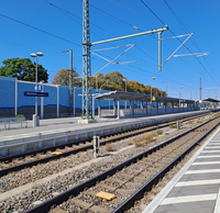 Der Hauptbahnhof Mülheim bei sonnigem Wetter mit mehreren Gleisen, einem Bahnsteig samt Sitzgelegenheiten, Lampenmasten, Überdachungen und elektrischen Oberleitungen.