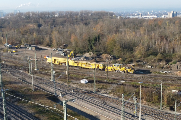 Ein gelber Bauzug auf Gleisen, im HIntergrund Wald und eine Stadt