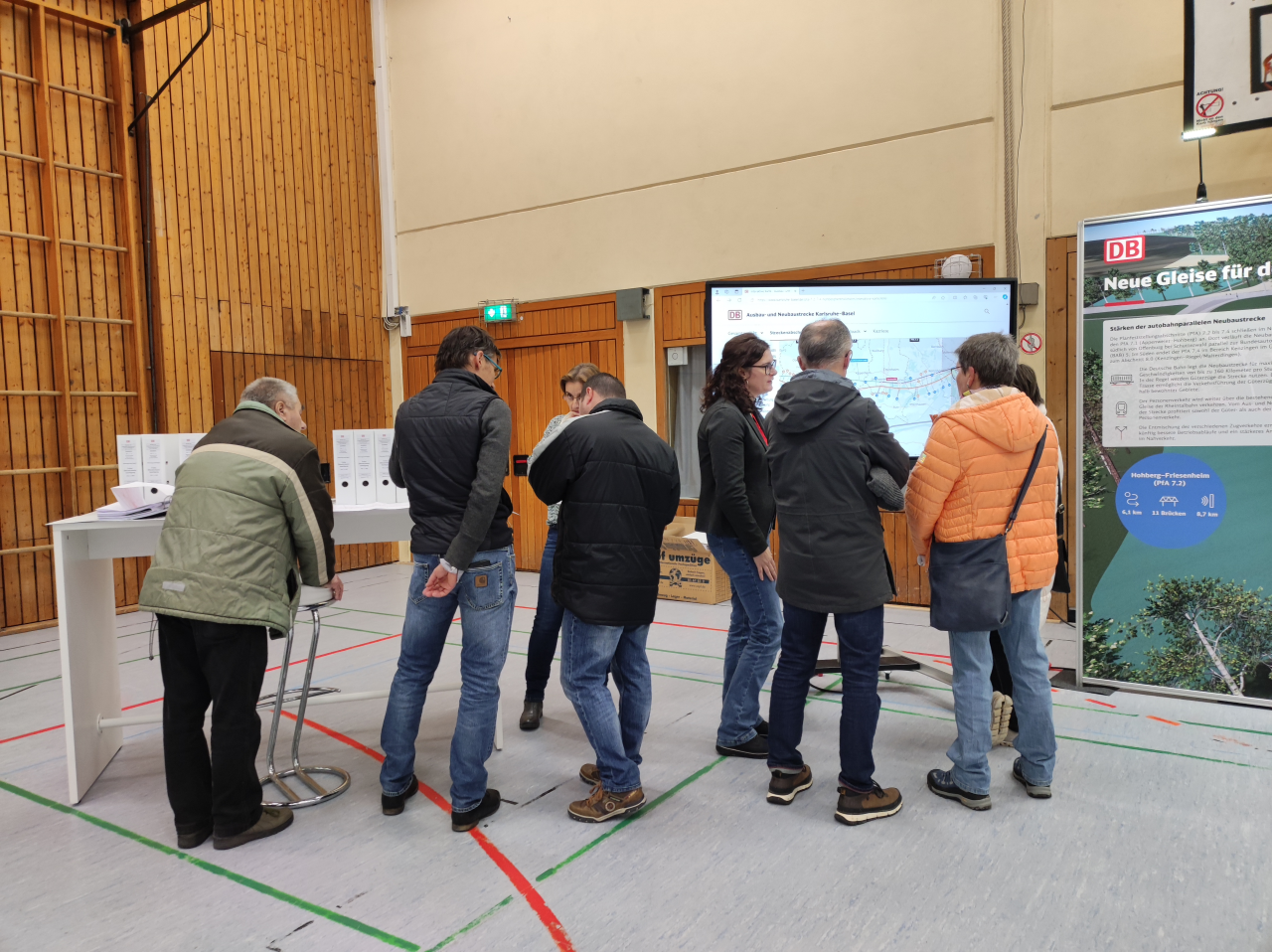 Mensch in einer Turnhalle an Infostand mit Tisch, Bildschirm und Messewand