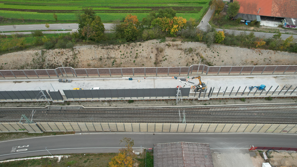 Eine Baustelle an einem Gleis. Ein Kran hebt Wandelemente zwischen Pfosten.