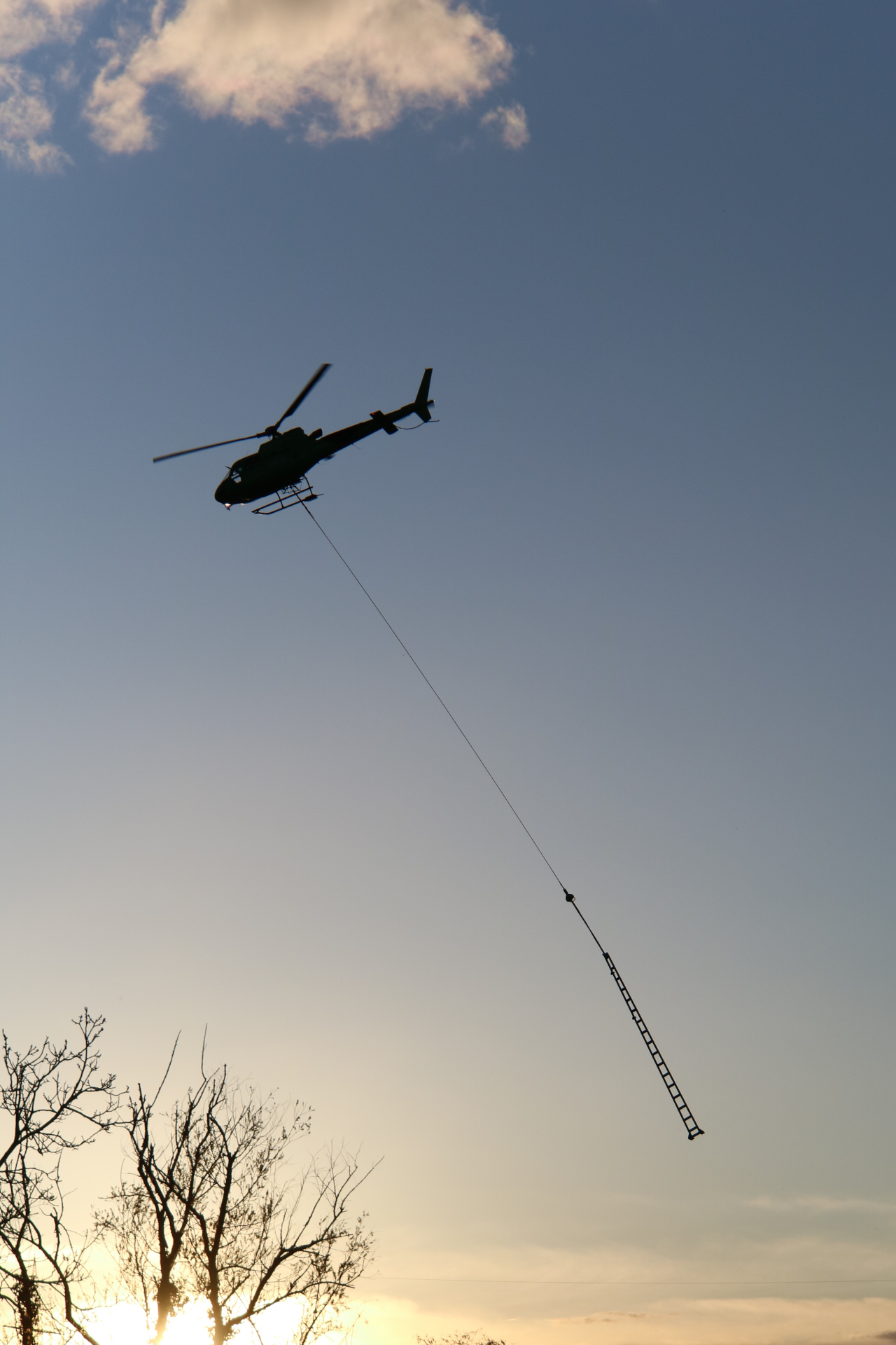 Ein Helikopter transportiert einen Oberleitungsmast bei Sonnenuntergang.
